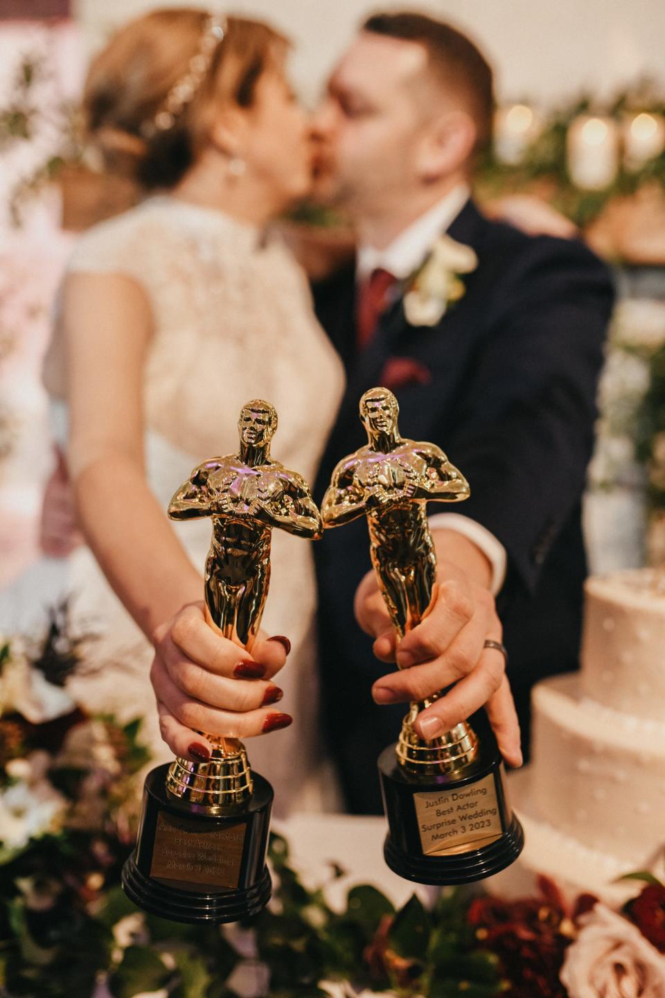 A bride and groom hold up trophies as they kiss.