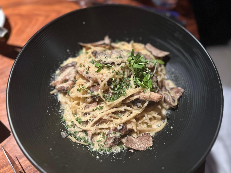 A black bowl of tagliatelle with pieces of mushrooms and truffle pieces with pieces of herbs on top