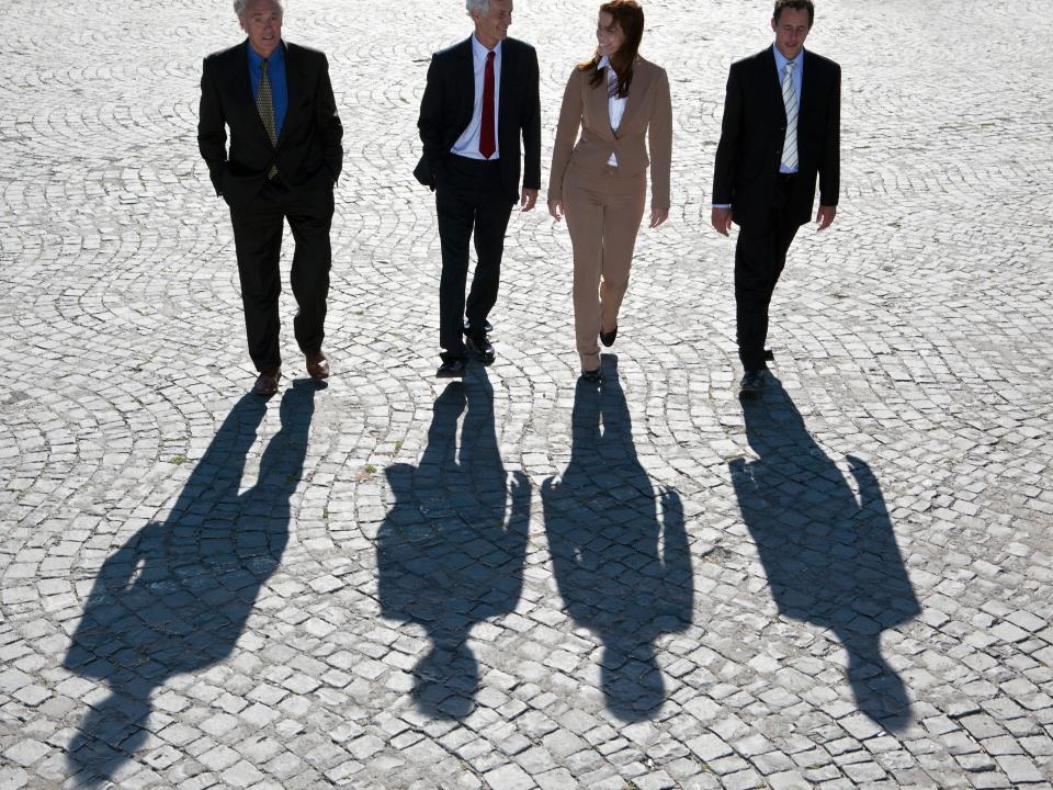 Four businesspeople wearing suits walk in a row along a cobbled street.