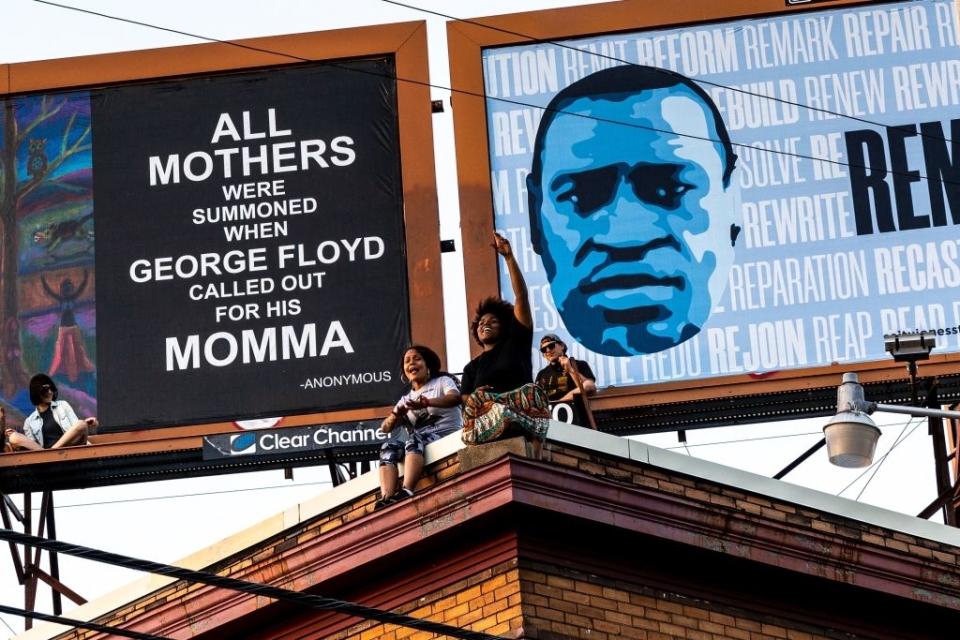 Protesters hold signs on a rooftop under billboards. One billboard has George Floyd's face; the other reads "ALL MOTHERS WERE SUMMONED WHEN GEORGE FLOYD CALLED OUT FOR HIS MOMMA"