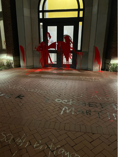 A brick building's doors and some of its stone facade covered in red paint. What appears to be spray-painted writing on the brick walkway makes a disparaging remark against weapons manufacturer Lockheed Martin.