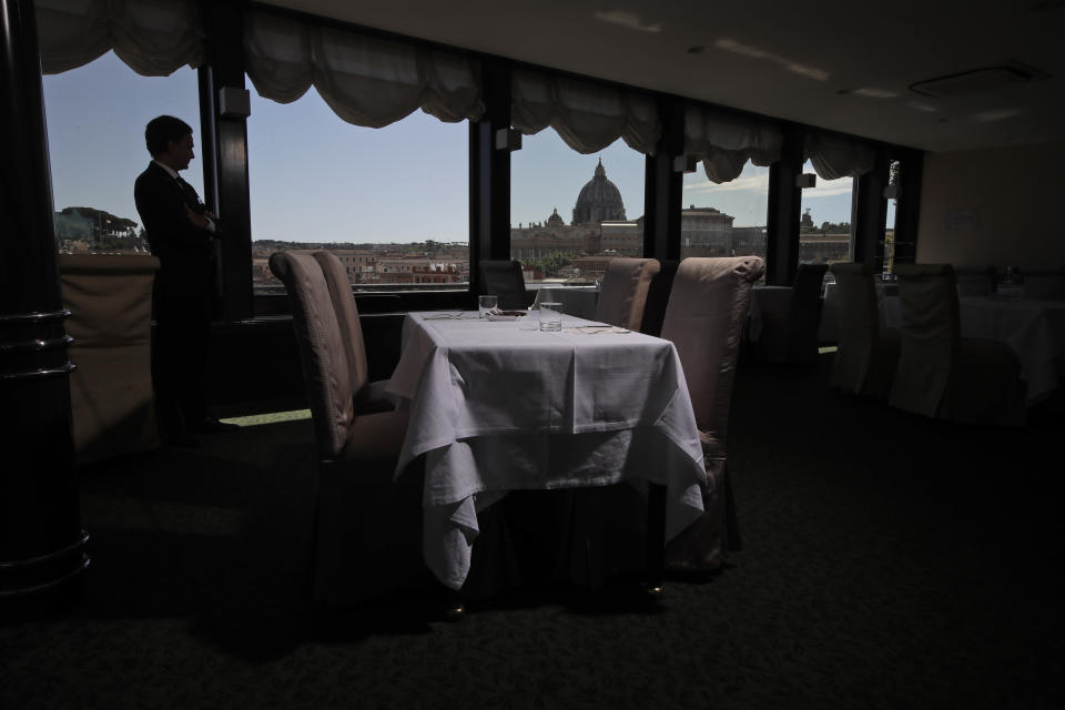 In this Monday, May 11, 2020 photo, Matteo Vasselli, concierge of Atlante Star Hotel, is silhouetted as he shows the restaurant of the hotel overlooking St. Peter's Dome, seen in the background, in Rome. The hotel remained open during the lockdown measures due to COVID-19, but has very few if no guests at all. (AP Photo/Alessandra Tarantino)