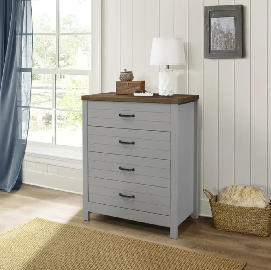 A gray four-drawer dresser with a brown top, a lamp, and decorative items in a room beside a window