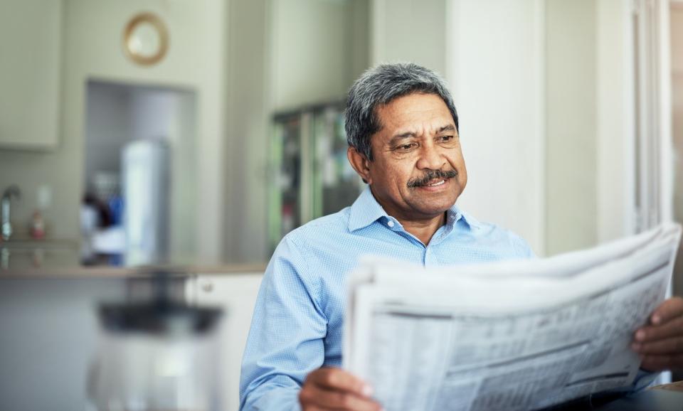 A smiling person reading a financial newspaper while seated in their home.