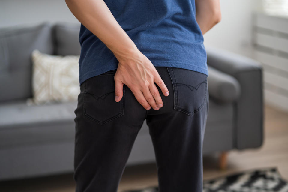 A person wearing a blue shirt and black pants is seen from the back, scratching their buttocks in a living room