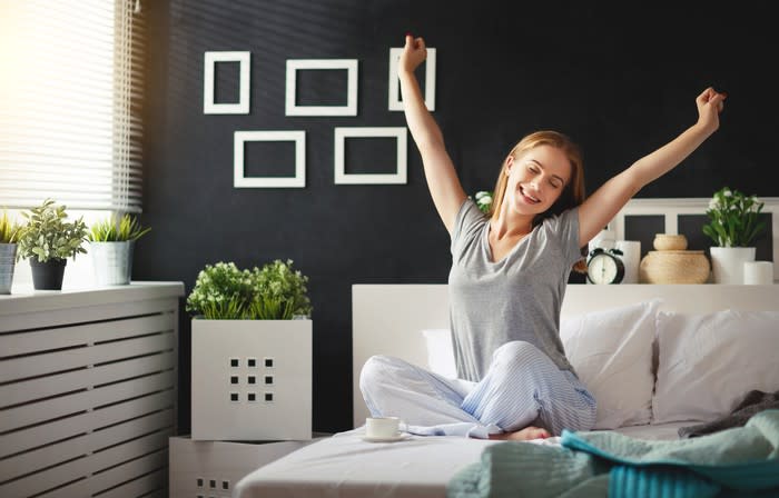 A smiling woman sitting cross-legged on a bed, stretching her arms above her head.