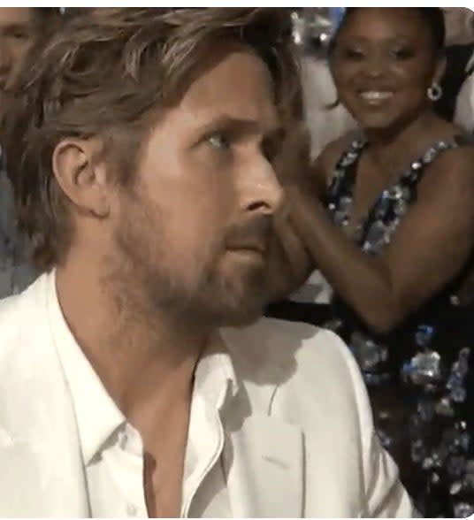 Ryan Gosling in a white suit, sitting and looking to his right with a focused expression. A woman in a sequined dress claps behind him