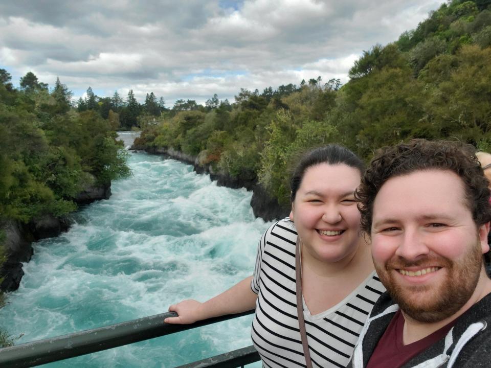 Riana Ang-Canning and her husband smiling in front of a blue body of water and green, lush trees in New Zealand