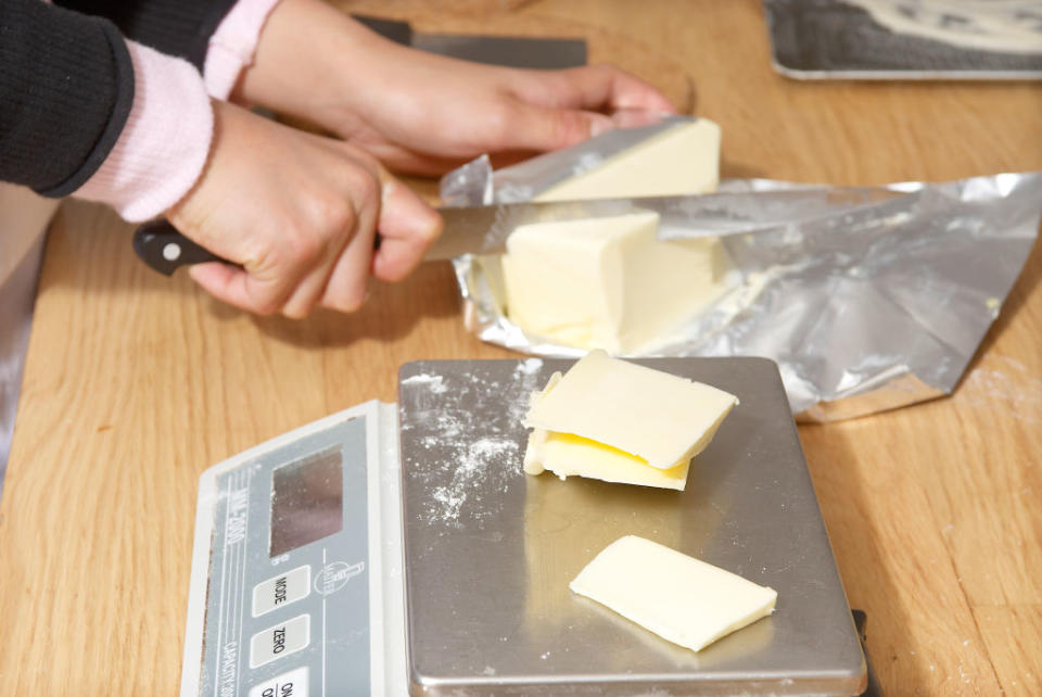 Hands cutting butter on a kitchen scale. The butter is partially wrapped in foil, and the person is using a knife to cut the butter into smaller pieces