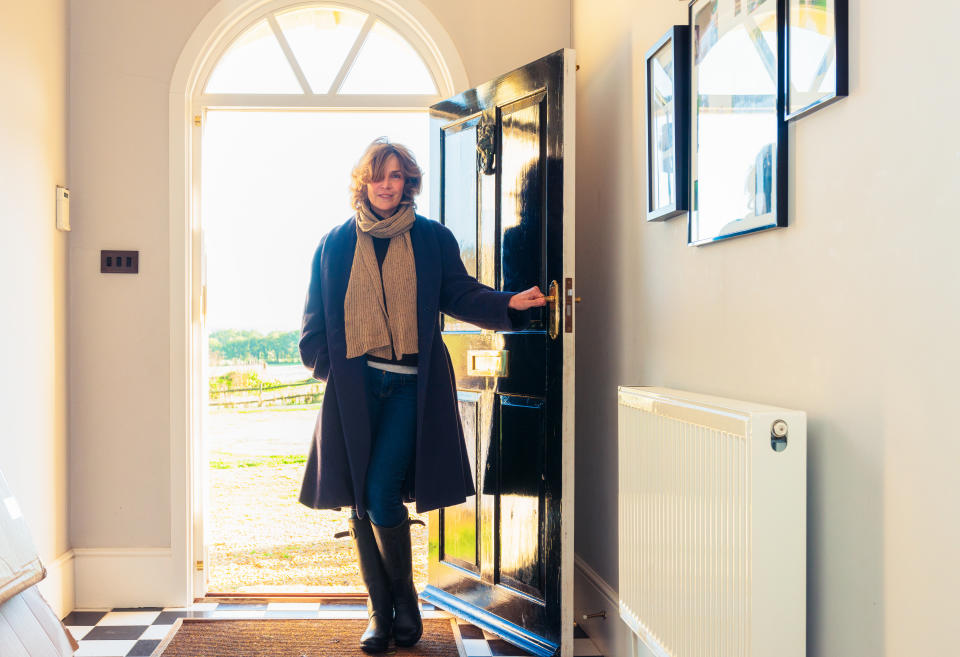 A woman holding her front door open, having returned home from a walk in the British countryside.