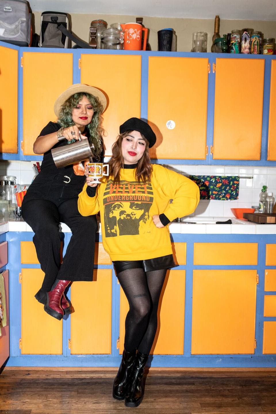 The two sisters pose in the kitchen, one pouring a kettle into a cup.