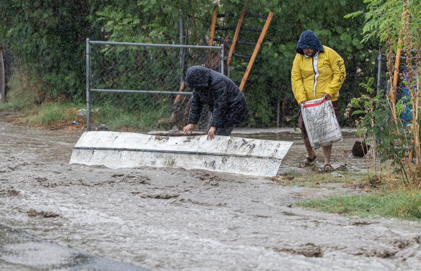 THERMAL, CA - AUGUST 20, 2023: Rafael Nunez, 29, right, with Isabel Ramirez, 23, carries bags of fertilizer to prop up a piece of roofing to try and divert flood waters on 70th Avenue from coming into his mobile home as tropical storm Hilary dumps torrential rain on the area on August 20, 2023 in Thermal, California. (Gina Ferazzi / Los Angeles Times)