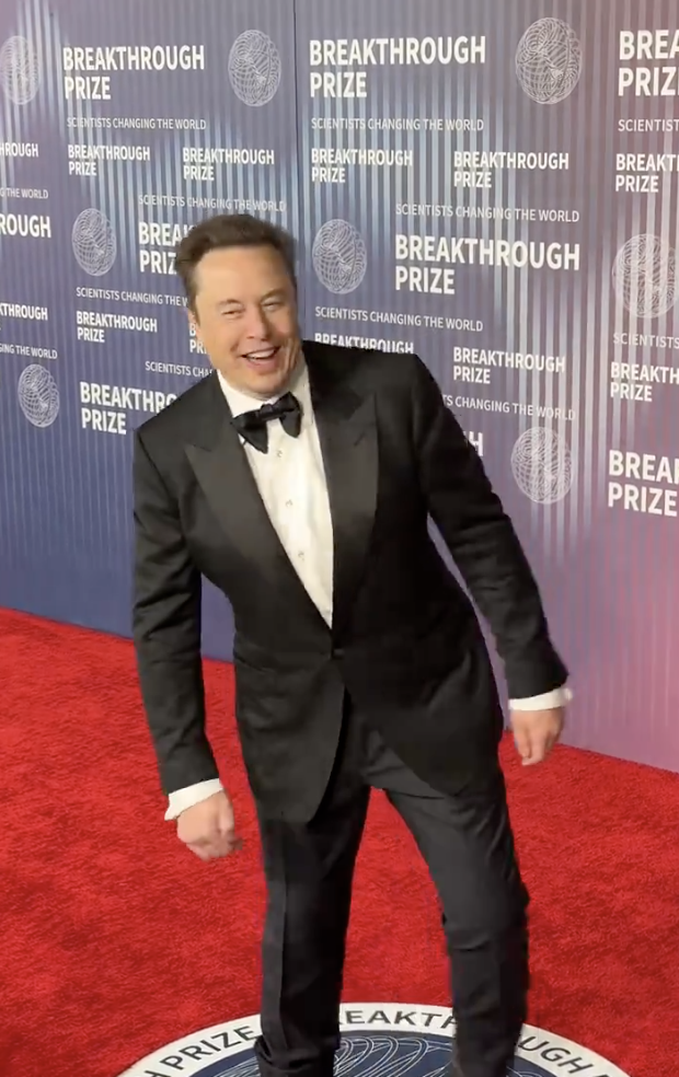 Elon Musk in a tuxedo with bow tie, smiling and gesturing thumbs-up on the Breakthrough Prize red carpet