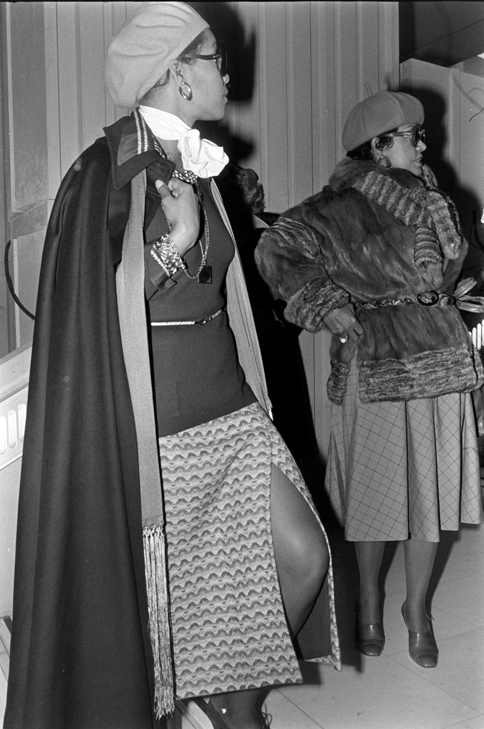 Black and white photo of Audrey Smaltz and Eunice Johnson dressed in cape, fur jacket and beret hats at a Paris fashion show in 1975