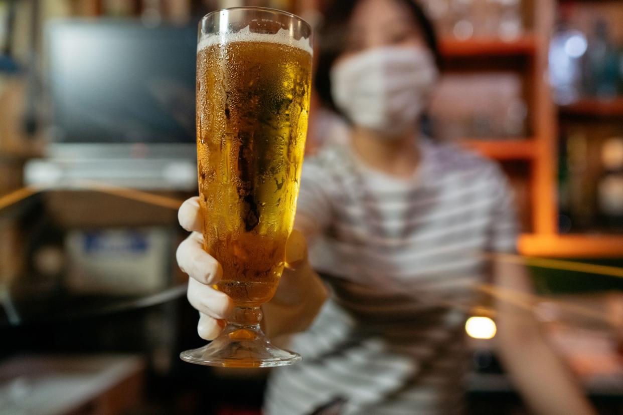 A bar owner is serving beer from behind a plastic clear curtain and she is wearing protective gloves and protective face mask to avoid illness spread.