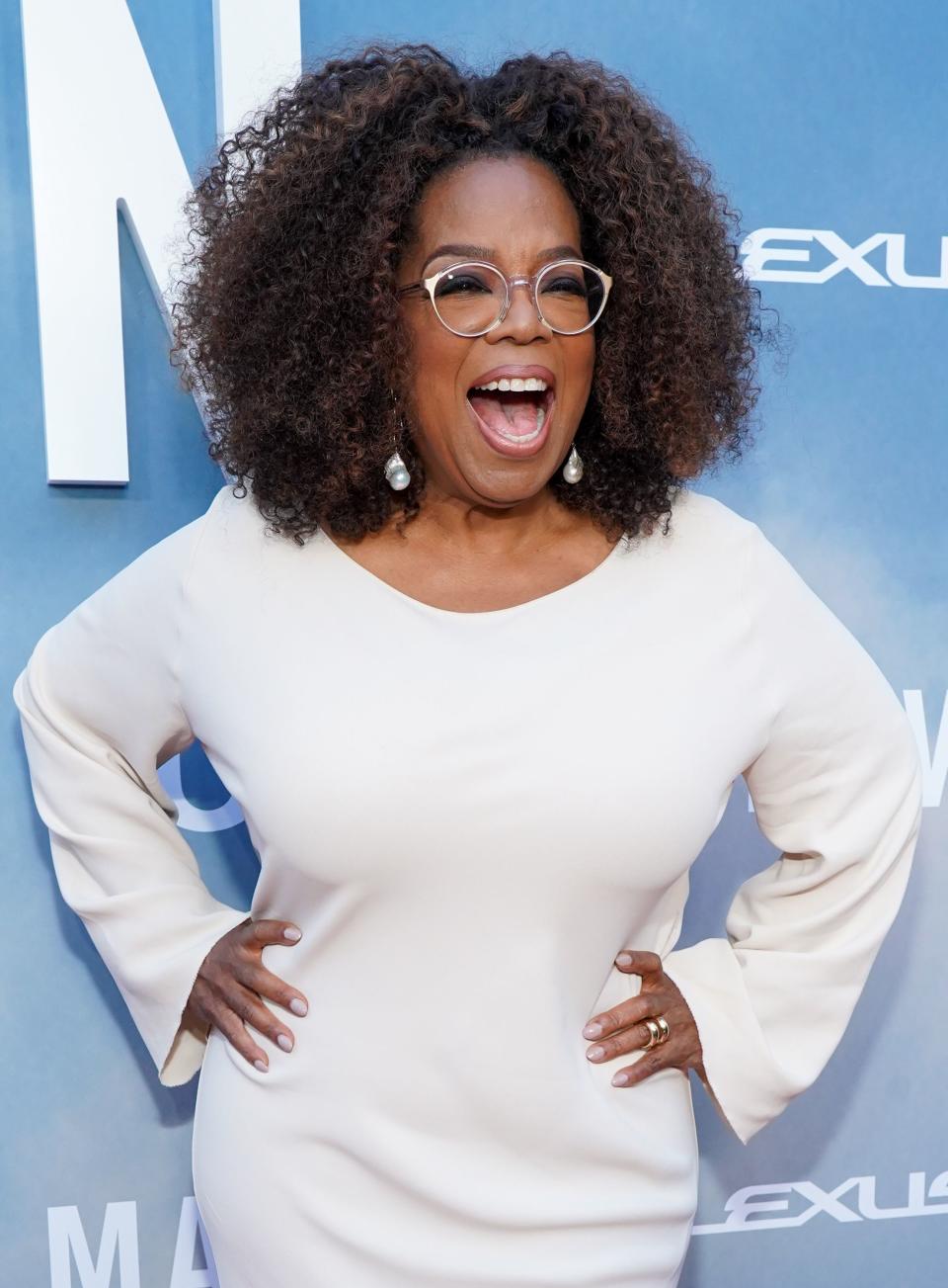 Oprah Winfrey stuns in white long-sleeve dress as she poses for the camera with her hands on her lips and a wide smile on her face. 