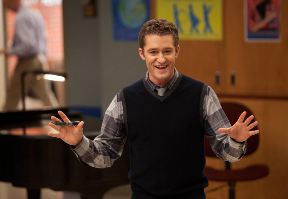 Actor in character on TV show set, wearing sweater over shirt with spread hands, expressing surprise or joy
