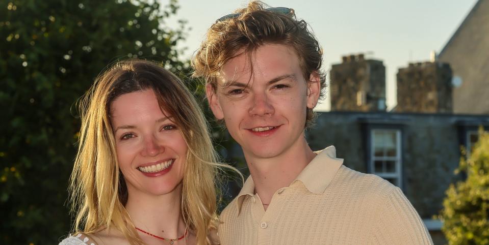 talulah riley and thomas brodie sangster, a young man and woman stand hugging as they smile at the camera, she has blonde hair worn down and wears a white dress, he has light brown curly hair and wears a beige top and trousers