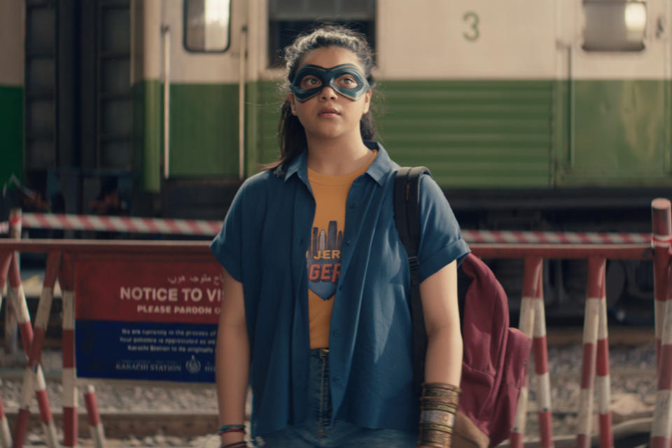 A teen girl in an orange tee and blue shirt, wearing a matching blue mask over her eyes, stands outside a train in Pakistan; still from "Ms. Marvel"
