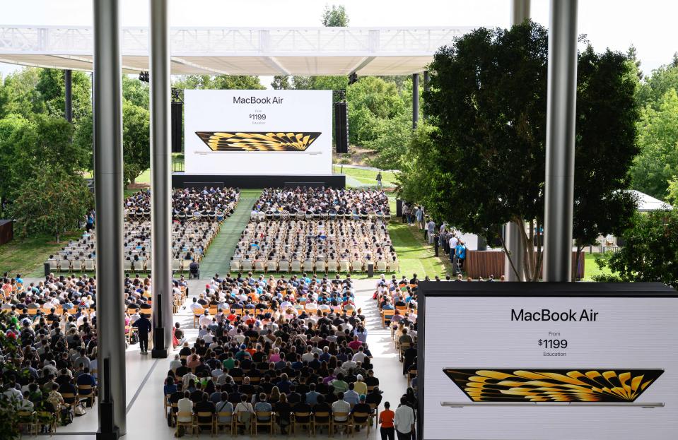 A crowd of people sitting in rows of chairs assembled on a large lawn as two large video screens show images of the MacBook Air on a white background.