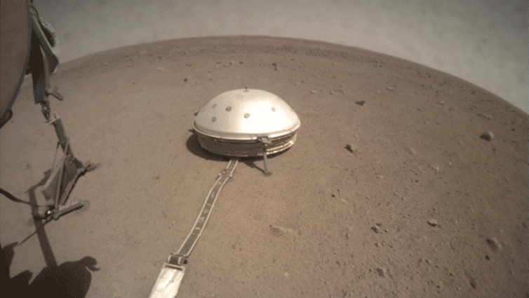 <span class="caption">Seen from InSight’s fisheye camera, its seismometer undisturbed on the martian surface under its dome-like wind shield, while nearby the robotic arm adjusts the digging instrument.</span> <span class="attribution"><span class="source">NASA/JPL-Caltech</span></span>