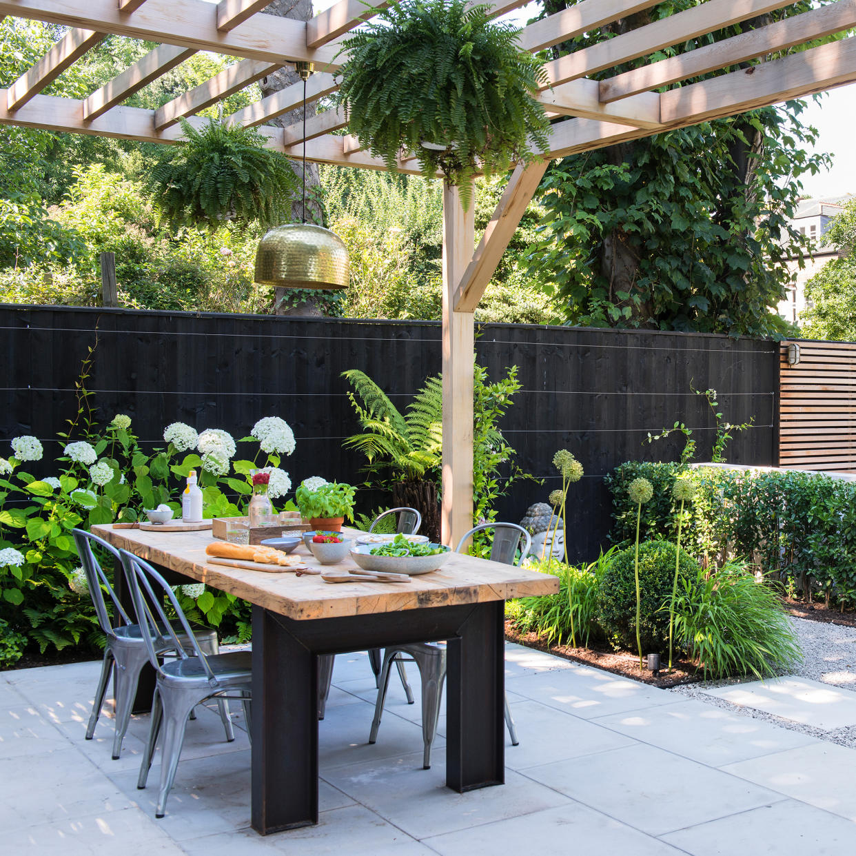  Patio with dining table with black legs and wooden top, metal bistro style chairs and a gold pendant light hung above from a wooden pergola. 