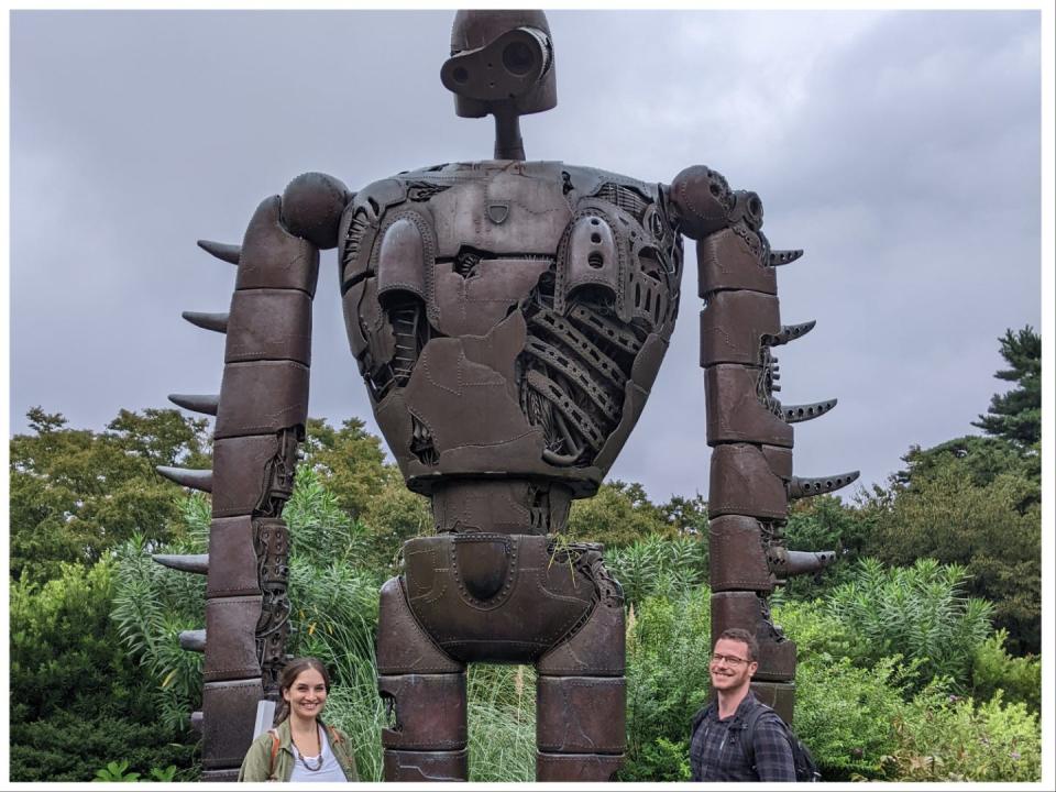 Image of India Kushner with her husband Sam LeGrys, smiling in front of a bronze robot statue that appears in the movie "Castle in the Sky." India has dark brown hair pulled back and wears a blue striped t-shirt and green jacket. Sam has short dark brown curly hair and wears a purple and black plaid shirt.