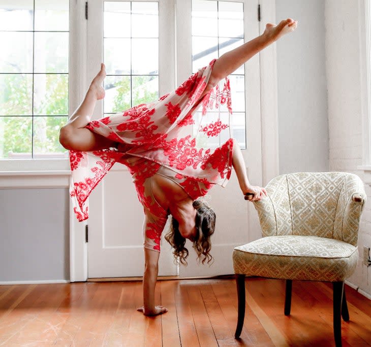 A woman practices Handstand in her living room, with one hand on a chair and one on the floor