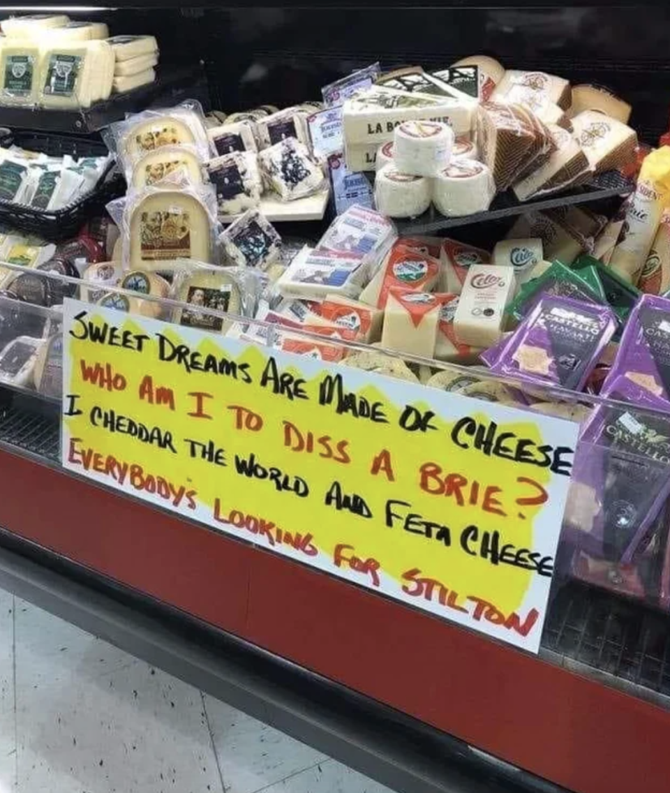 Handwritten signs by a cheese display: "Sweet dreams are made of cheese / Who am I to diss a Brie? / I cheddar the world and feta cheese / Everybody's looking for Stilton"