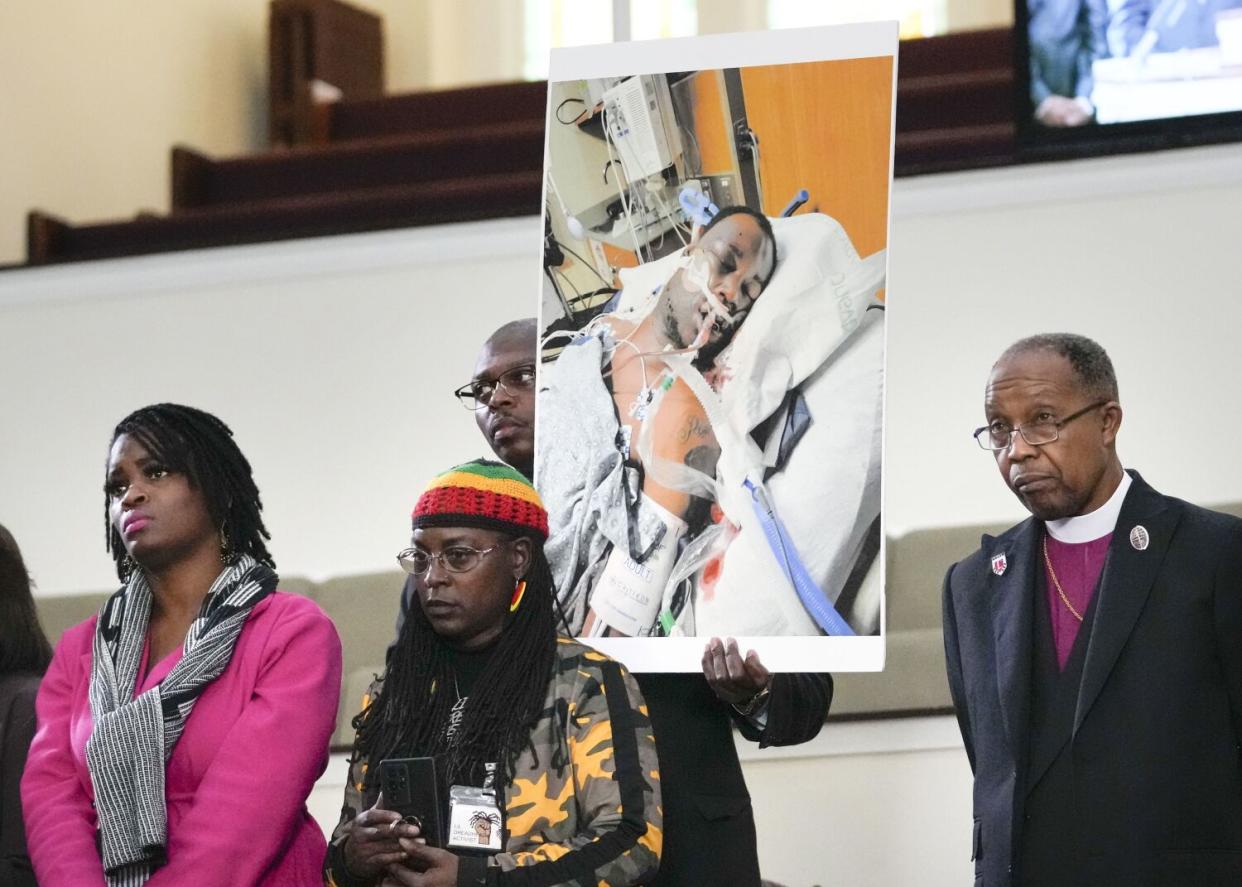 A man with a poster of an injured man in a hospital bed, standing somberly with others, including a clergyman.