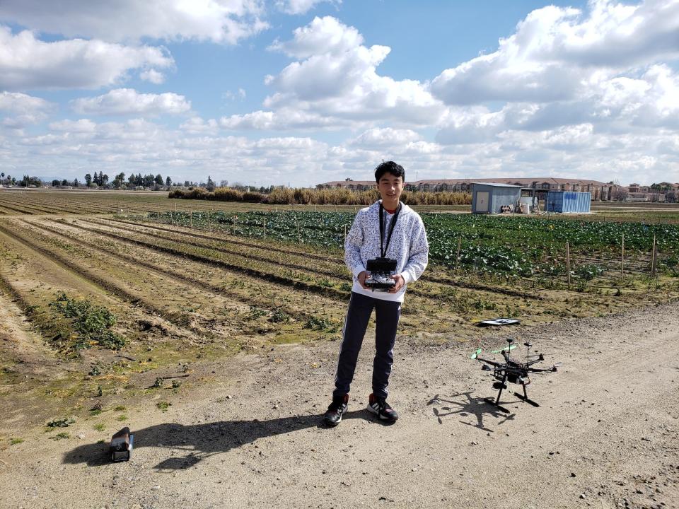 teenage boy standing in a crop field holding the controller for a drone on the dirt path next to him