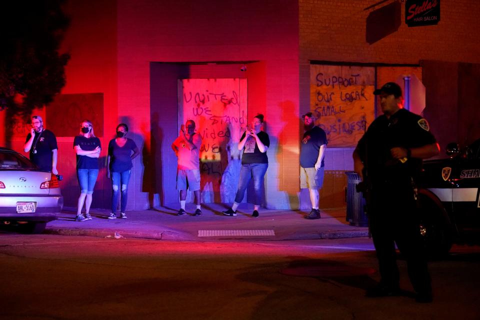 Bystanders watch outside boarded-up shops as protesters clash with police late Tuesday, Aug. 25, 2020, in Kenosha, Wis.