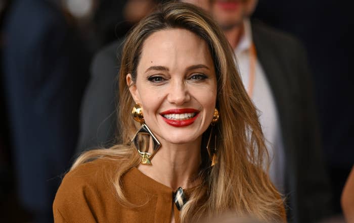 Angelina Jolie smiles while wearing geometric earrings and a brown top at a public event