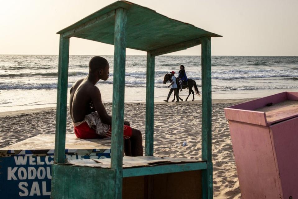 A young man looks as a woman takes a horse ride along a beach in Banjul.