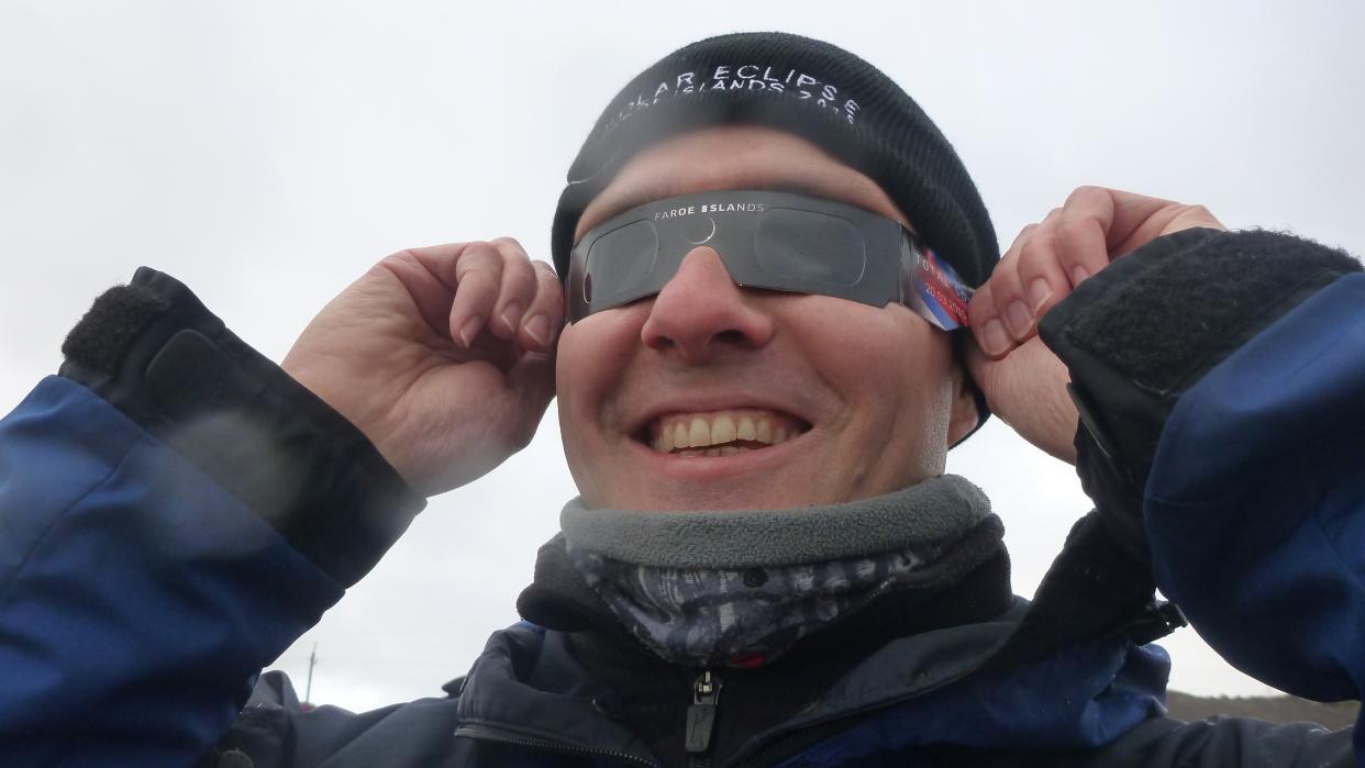  A man wearing eclipse glasses and a hat, he is smiling. . 