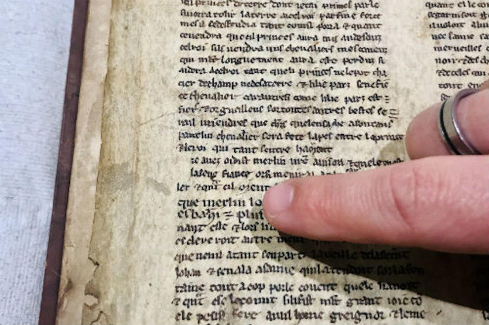 A finger points to the name "Merlin" on a page with text in a very old book