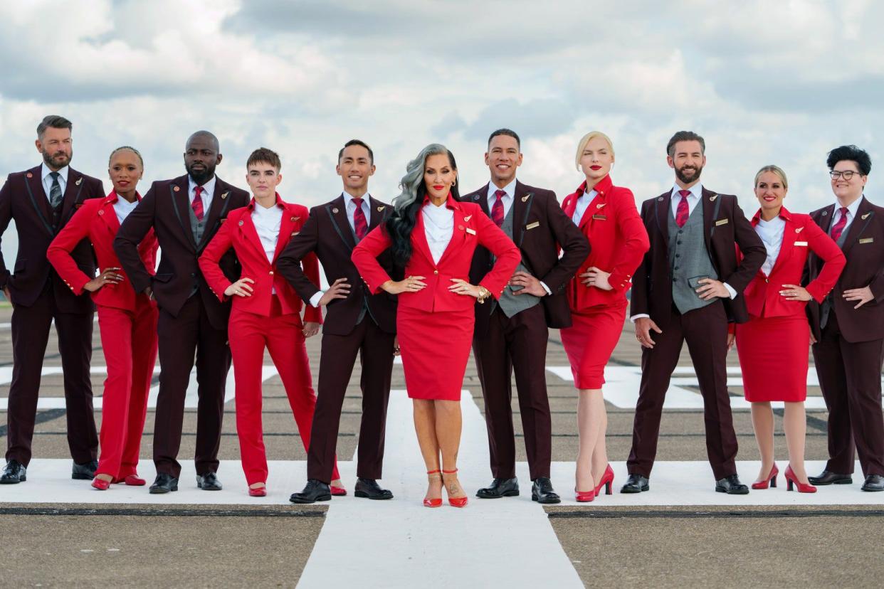 Eleven airline stewards and stewardesses showcasing alternating brown and red Virgin Atlantic uniforms in different style combinations, dramatically, on a white runway outside, with clouds in the sky in the background