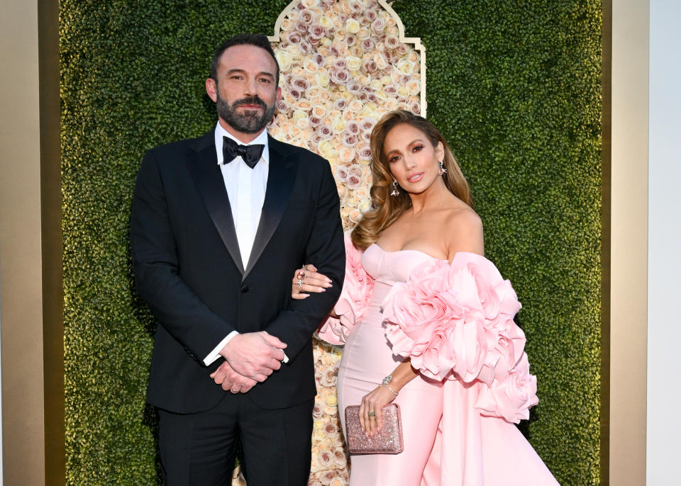 Ben Affleck and Jennifer Lopez at a formal event posing in front of a floral backdrop. Ben wears a tuxedo, and Jennifer wears an elegant gown with ruffled sleeves