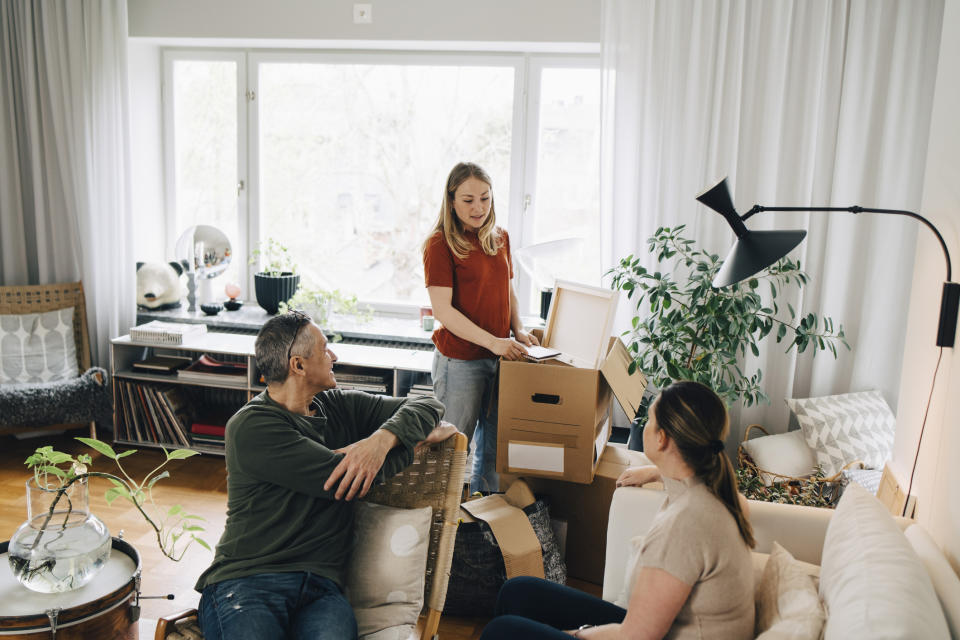 A discussion around unpacked boxes in a living room, involving three people seemingly planning or organizing, associated with work and moving topics