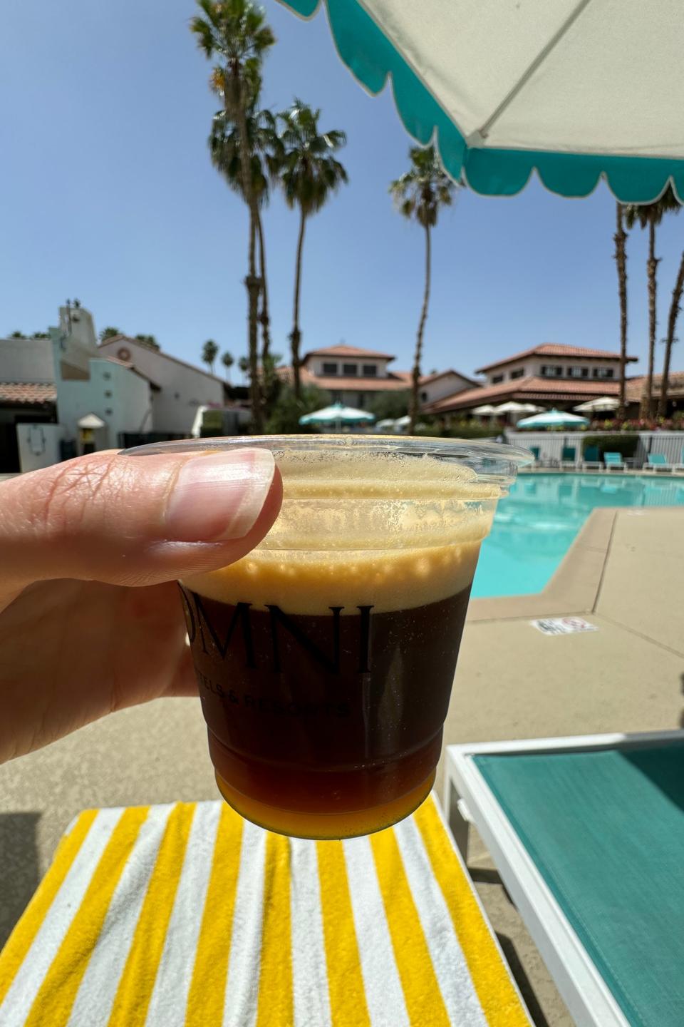 Person holding a cup of coffee by a pool with palm trees and loungers in the background
