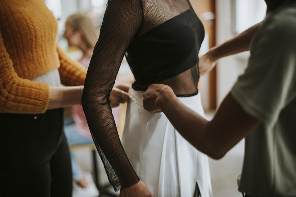 Person in a sheer black top being assisted into a white skirt by two individuals