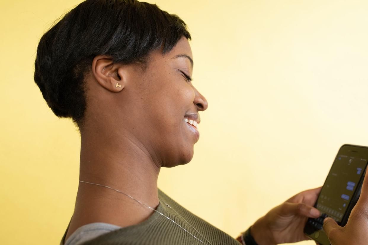 Reference this list of conversation starters for texting when you want to talk to someone. Pictured: A Black woman standing to the side and smiling at text messages on her phone.