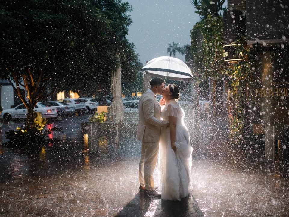 A bride and groom kiss on a street under an umbrella surrounded by rain.