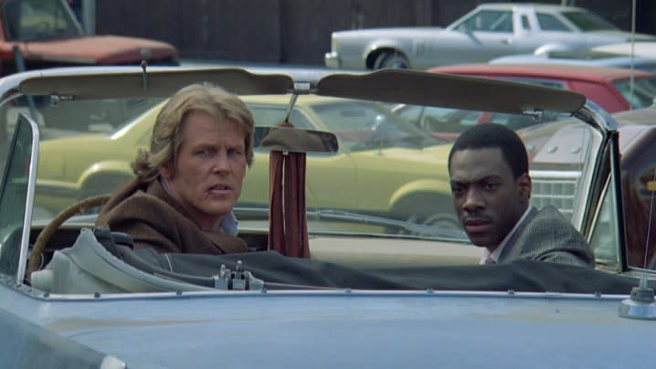 Nick Nolte and Eddie Murphy sit in a car and look backwards.