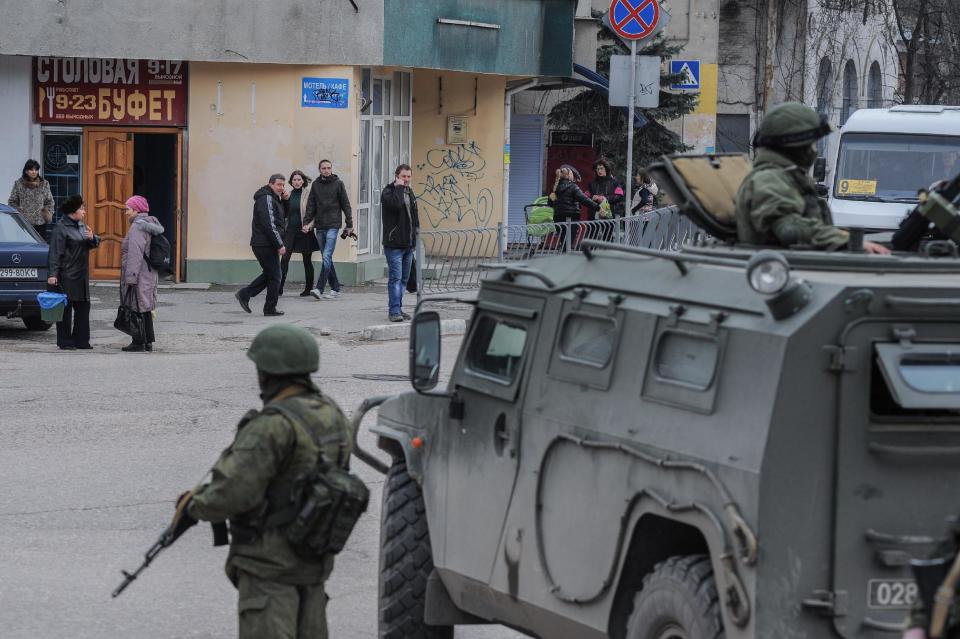 Troops in unmarked uniforms stand guard in Balaklava as people walk in a street, on the outskirts of Sevastopol, Ukraine, Saturday, March 1, 2014. An emblem on one of the vehicles and their number plates identify them as belonging to the Russian military. Ukrainian officials have accused Russia of sending new troops into Crimea, a strategic Russia-speaking region that hosts a major Russian navy base. The Kremlin hasn’t responded to the accusations, but Russian lawmakers urged President Putin to act to protect Russians in Crimea. (AP Photo/Andrew Lubimov)