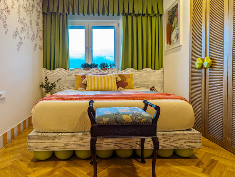 Bedroom with tree branch wallpaper wall, green curtains, parquet wood floor, and pops of yellow and orange