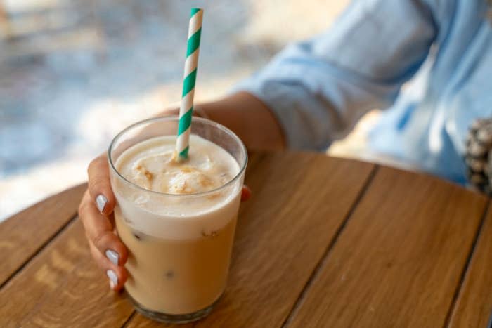 A person holds a glass of iced coffee with a green and white striped straw on a wooden table