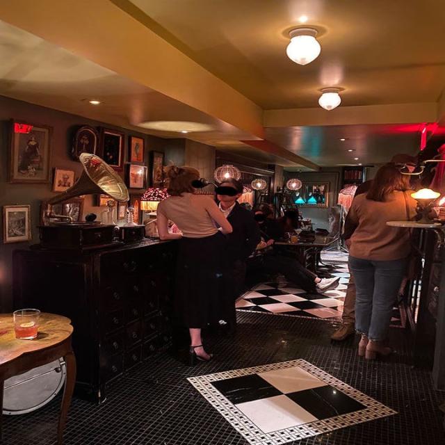People standing and sitting in the speakeasy, with checkerboard flooring, and a gramophone on the bar