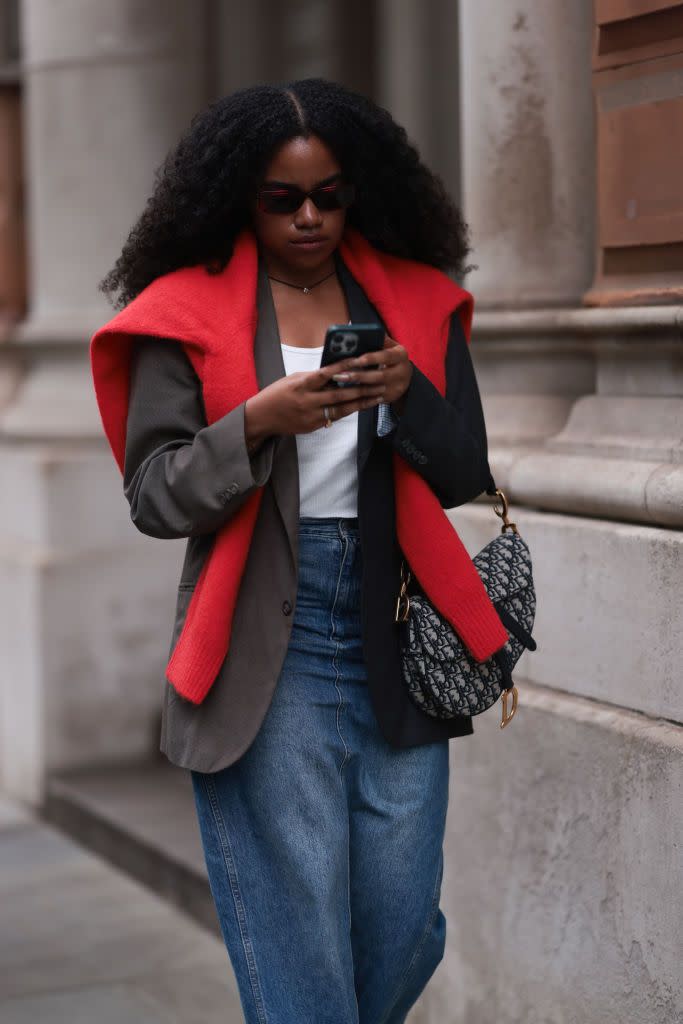 A girl at London Fashion Week is wearing a blazer and red jumper, on top of which she wears mid-blue jeans and a Christian Dior bag, on the phone