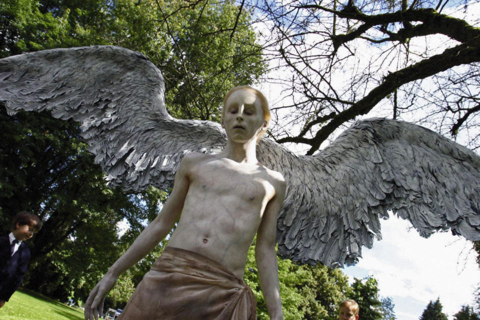 A pale young man with angelic wings stands among children in the sun